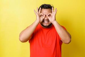 Funny cheerful guy holds hands near eyes, pretends looking through binoculars tries to find love of life, wearing red t-shirt, shows zero or okay gesture over eyes, isolated on yellow background photo