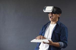 Young man in casual style using Virtual Reality glasses and clicking wireless keyboard on dark grey background. Copy space for futuristic concept. photo