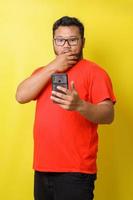 Fat asian guy with surprised expression covering mouth while looking to cell phone isolated on yellow background photo
