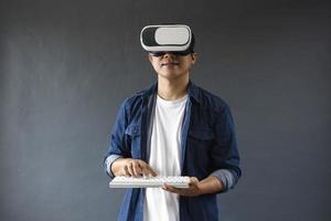 Young man in casual style using Virtual Reality glasses and clicking wireless keyboard on dark grey background. Copy space for futuristic concept. photo
