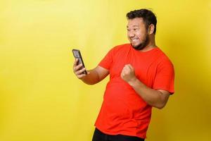 Excited fat asian man with celebrating gesture while holding mobile phone isolated on yellow background photo