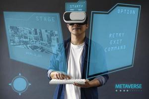 Asian happy man starting the games with hologram screen using VR googles and wireless keyboard. Virtual games futuristic concept, dark grey background. photo