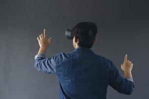 Back view of young man in casual style wearing virtual reality googles, his finger pointing out or touching blank screen. Copy space for futuristic games and education concept. photo
