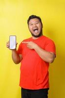 Attractive asian fat man in red tshirt is pointing finger to blank screen smartphone isolated on yellow background photo