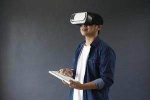 Young man in casual style using Virtual Reality glasses and wireless keyboard on dark grey background. Copy space for futuristic concept. photo