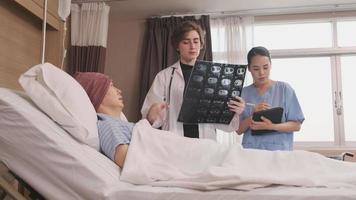 Caucasian female doctor in uniform diagnosis explains x-ray film with Asian radiologist and recovery male patient at inpatient room bed in a hospital ward, medical clinic, cancer examination consult. video