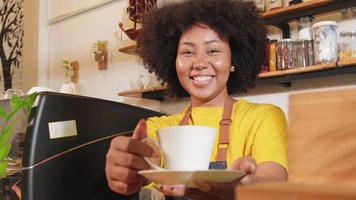 African American female barista in looks at camera, offers cup of coffee to customer with cheerful smile, happy service works in casual restaurant cafe, young small business startup entrepreneur. video