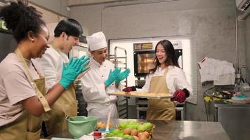 Junge asiatische Studentin der Kochklasse bringt ein Tablett mit gebackenen Kuchen aus dem Elektroofen, Seniorchef daneben, fröhliche Konditoreiküche im Kochkurs, Lebensmittelbesetzung in der Edelstahlküche. video