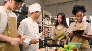 Hobby cuisine course, senior male chef in cook uniform teaches young cooking class students to peel and chop apples, ingredients for pastry foods, fruit pies in restaurant stainless steel kitchen. video