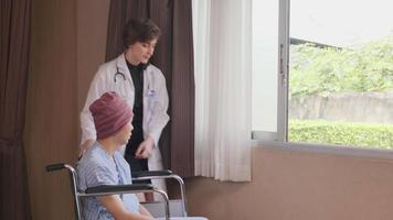 une jeune femme médecin de thérapie caucasienne en uniforme pousse un patient masculin en fauteuil roulant vers la fenêtre, soutient et motive la guérison d'une maladie cancéreuse après un traitement médical de chimio dans une chambre d'hôpital. video