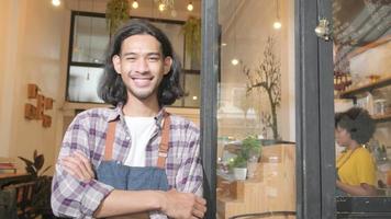 A long hair handsome Asian male startup barista with apron standing at casual cafe door, arms crossed, looking at the camera with and welcoming smile, happy and cheerful with coffee shop service jobs.