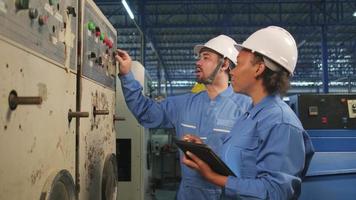Professional industry engineer teams in hard hats and safety uniforms inspect machine's control panel, maintenance check with tablet in mechanical manufacture factory, electrical service occupation.