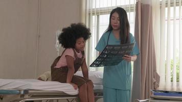 Asian female doctor in uniform health checks African American child, illness patient, diagnosis explains x-ray film in emergency room bed at hospital ward, pediatric clinic, kids examination consult. video
