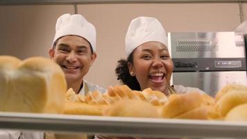 retrato de chefs profissionais de uniforme branco olhando para a câmera com um sorriso alegre e orgulhoso com bandeja de pão na cozinha. um amigo e parceiro de alimentos de panificação e ocupação diária de padaria fresca. video