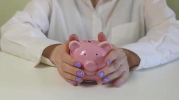 Close-up of female hands, puts coins in a pink piggy bank on a wooden table. video