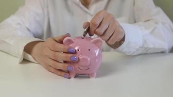 Close-up of female hands, puts coins in a pink piggy bank on a wooden table. video