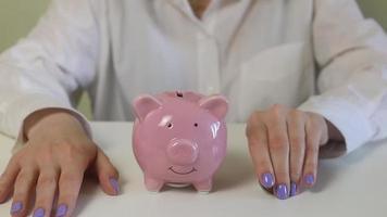 Close-up of female hands, puts coins in a pink piggy bank on a wooden table. video