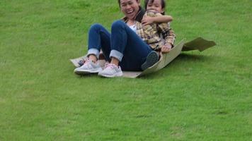 Lachende junge Mutter und kleines Mädchen sitzen auf einem Karton, der im Garten einen Hügel hinunterrutscht. Konzept der glücklichen Kindheit.