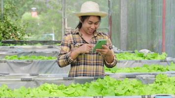 Dona de jardineira asiática recebendo pedidos on-line perto de viveiro de alfaces jovens em fazenda orgânica com felicidade. agricultora usando telefone celular na fazenda de vegetais orgânicos. video