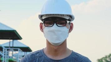 joven ingeniero asiático con casco y máscara mira y sonríe a la cámara en el fondo de la presa. video