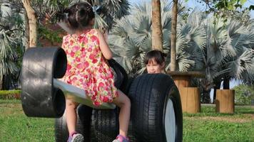 bambini carini che giocano nel parco giochi all'aperto. le sorelline si siedono su una sega fatta di vecchi pneumatici nel parco. sana attività estiva per i bambini. video