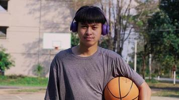 jonge aziatische atleet die een koptelefoon draagt, poseert met basketbal op een buitenbaan. video