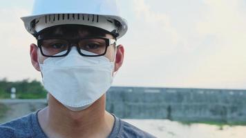 Young Asian engineer wearing a helmet and mask looks and smiles at the camera on the dam background. video