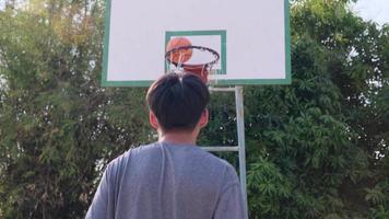 joven atleta asiático con auriculares posa con baloncesto en la cancha al aire libre. video