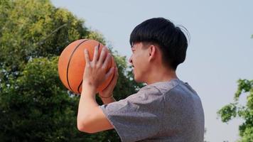 jovem atleta asiático usando fones de ouvido posa com basquete na quadra ao ar livre. video