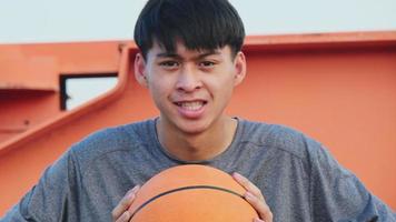 jonge aziatische atleet die een koptelefoon draagt, poseert met basketbal op een buitenbaan. video