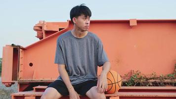 Young Asian athlete wearing headphones poses with basketball at outdoor court. video