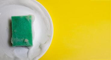 Green sponge in foam on a white plate on a yellow background. Washing dishes concept photo