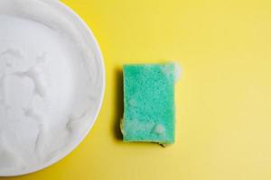 Green sponge in the foam and a white plate on a yellow background. Washing dishes concept photo