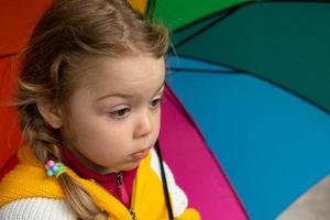 A 3-year-old girl hides in the rain under a colored umbrella photo