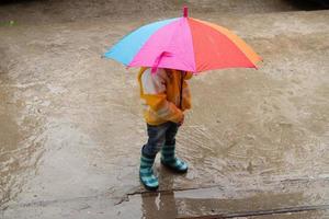 una niña de 3 años se esconde bajo la lluvia bajo un paraguas de colores foto