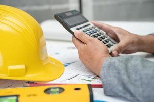 Asian engineer with architectural house plan project blueprint, graph and yellow helmet and engineering construction tools. photo