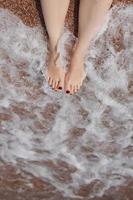 concepto de viaje - piernas de mujer en una hermosa playa tropical con arena de guijarros. pies sobre arena y olas en verano. chica descalza en el agua del océano en viajes de vacaciones. siéntete feliz y relájate. copie el espacio foto