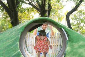 Cute little girl and young mother playing together in the mountains with tunnel in the outdoor playground. Healthy summer activity for children.