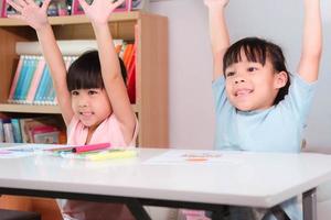 Happy kids with arms up sit in classroom. Cute girl taking lessons for home schooling Elementary school children enjoy learning together. Back to school concept. photo