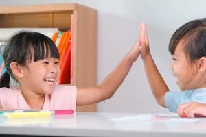 Little girl giving a hi-five with to her friends sitting in the classroom. Cute little sisters taking lessons for home schooling. Elementary school children enjoy learning together. Back to school photo