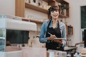 mujer feliz dueña del café de pie detrás del mostrador de una cafetería. barista femenina de pie y sosteniendo una tableta digital detrás del mostrador en la cafetería. concepto de propietario de negocio. foto