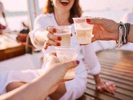 young women hands holding plastic glasses with sparkling wine during travelling on yacht. plastic, environment, recycling, toast, party concept photo