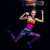 redhead funny woman in colorful clothes and pink sunglasses on the dark background swimming underwater with photo camera. Travel, sightseeing concept