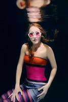 redhead funny woman in colorful clothes and pink sunglasses on the dark background swimming underwater photo