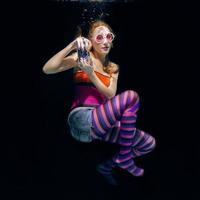 mujer pelirroja divertida con ropa colorida y gafas de sol rosas en el fondo oscuro nadando bajo el agua con cámara fotográfica. viajes, concepto de turismo foto