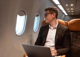 Businessman working with laptop while sitting in airplane seat and looking through aeroplane window. Business traveler concept photo