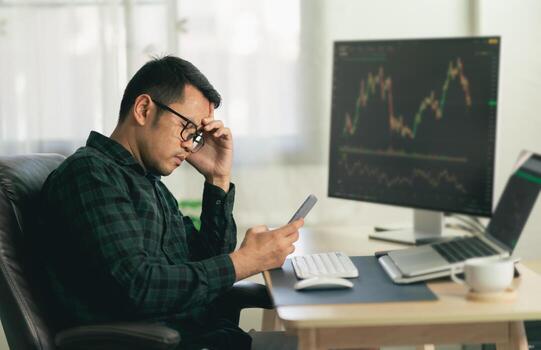 Asian man trader unhappy and serious sitting at home office in front of monitors with cryptocurrency graph holding smartphone monitoring cryptocurrency price photo