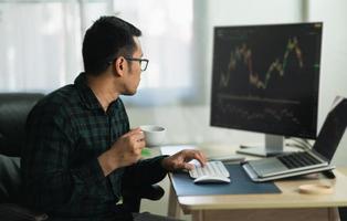 Asian man trader drinking coffee and sitting at home office in front of monitors with cryptocurrency graph holding smartphone monitoring cryptocurrency price photo