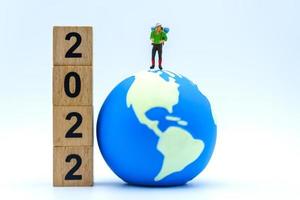 2022 año nuevo y concepto de viaje. Primer plano de figuras en miniatura de viajero con mochila de pie en una mini bola mundial con una pila de bloques de números de madera sobre fondo blanco. foto