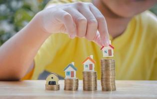 niño asiático que tiene una casa o una casa en una pila de monedas de oro para ahorrar dinero, invertir para el futuro y comprar una casa. concepto de préstamo, escalera de propiedad, inversión financiera, inmobiliaria y bonificación. foto